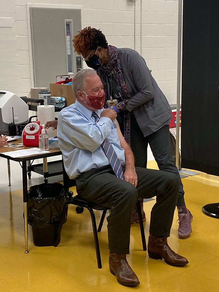 A photo of Superintendent Woods and a healthcare worker in a school setting.  The Superintendent is seated and smiling as the healthcare worker administers a vaccine to his shoulder. Both are wearing cloth masks.